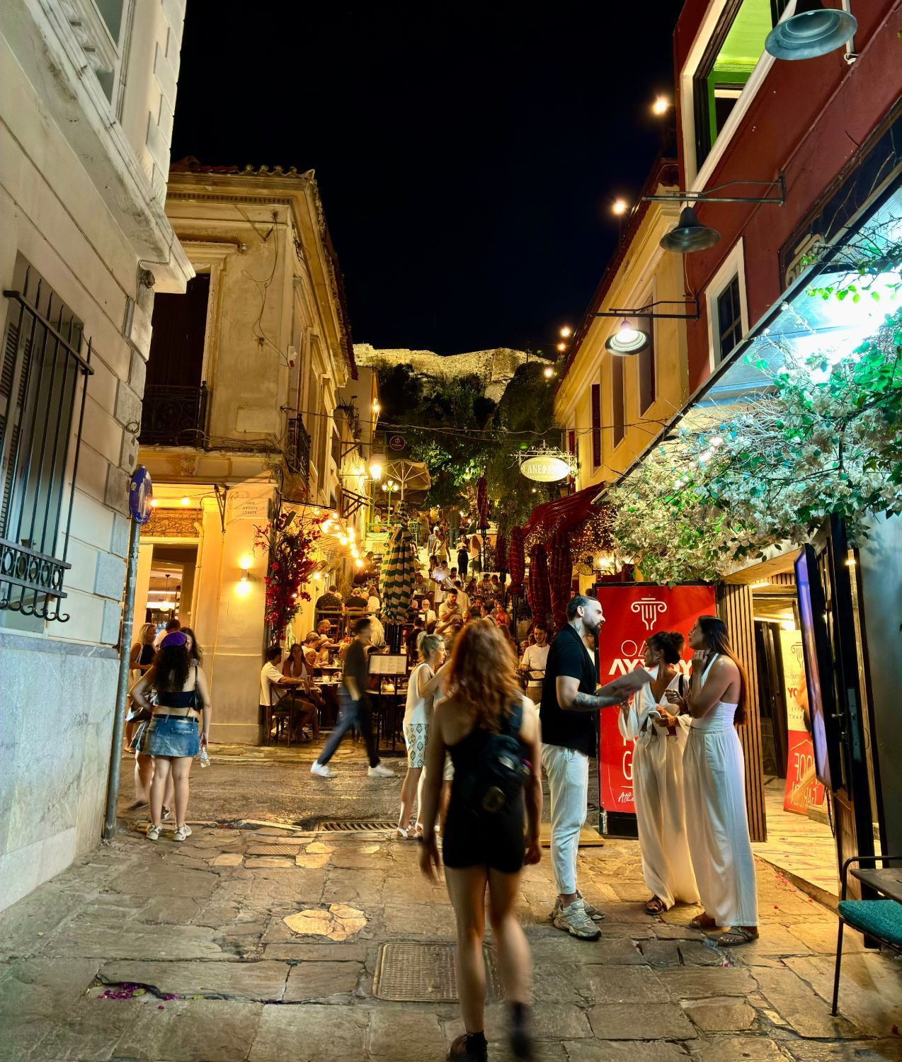 A busy stepped street in Plaka at night lined with restaurants, with the floodlit Acropolis above