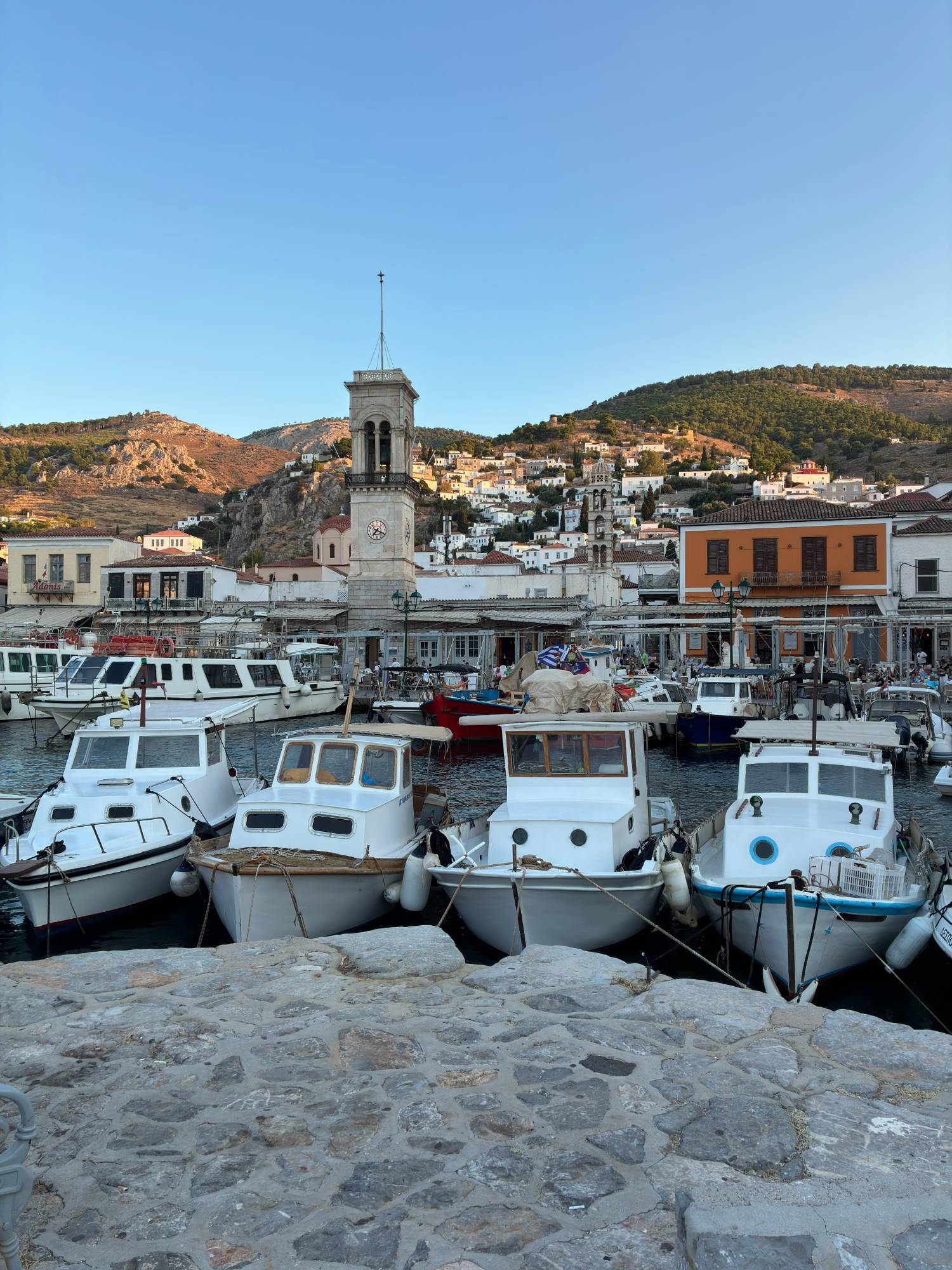 Fishing boats in Hydra harbour in front of the clock tower, with white houses climbing the hills