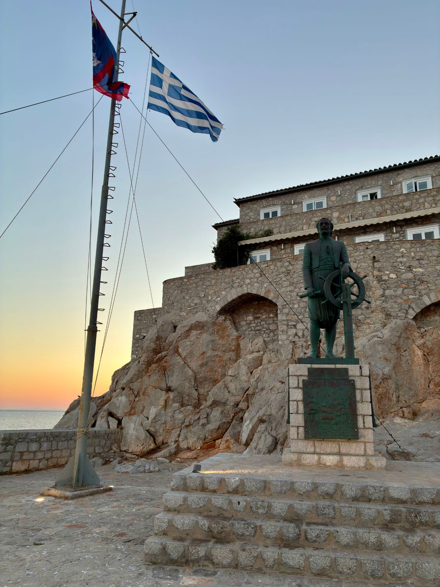 Bronze statue of a sea captain at a ship's wheel beside the Greek flag on the Hydra waterfront at dusk