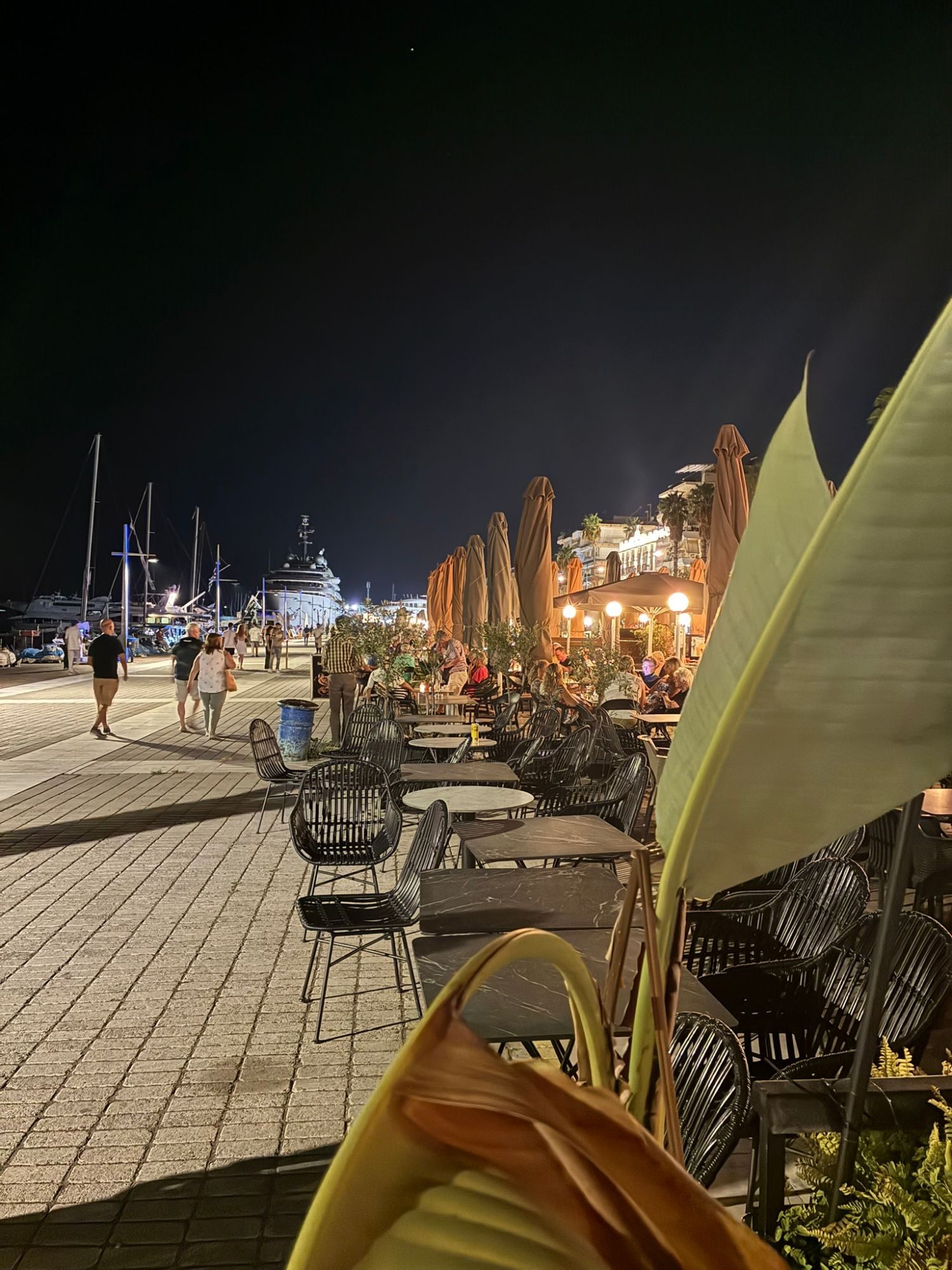 Kalamata's harbour promenade at night with café tables, people strolling and a superyacht in the background