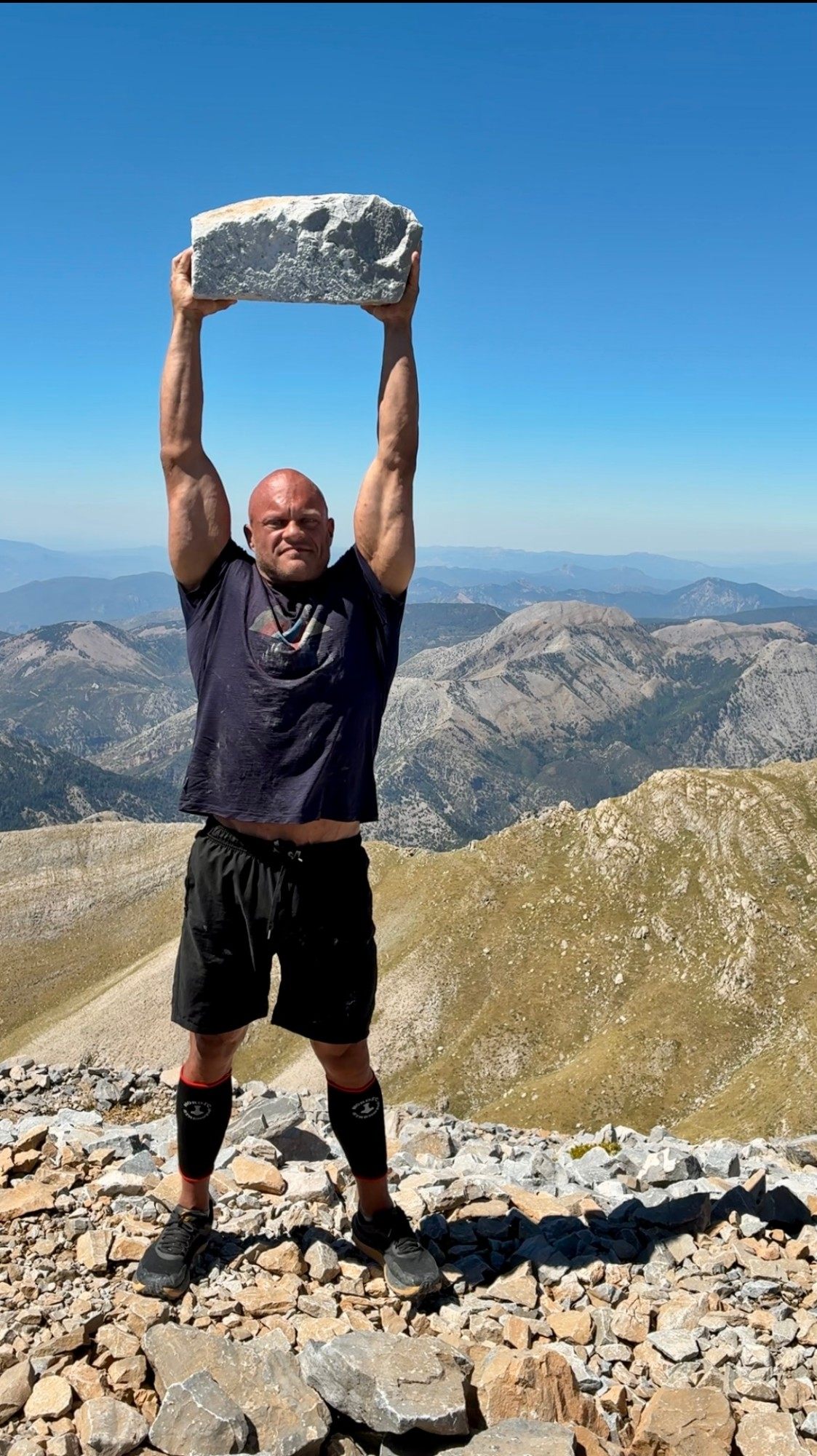 Lifting a large rock overhead on the summit of Mount Taygetos with mountain ridges behind