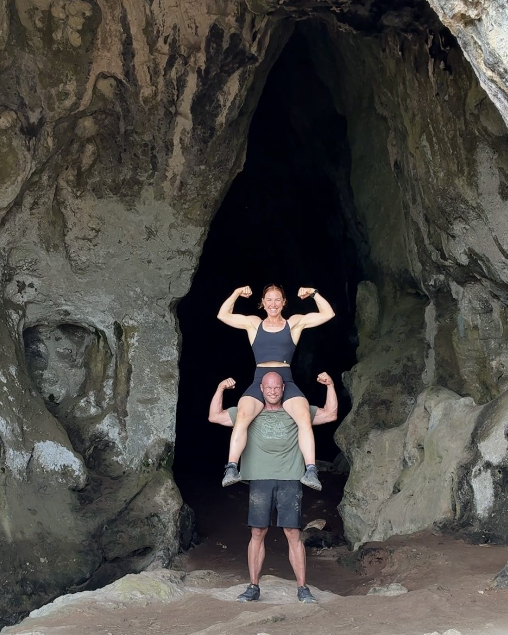 Kristine sitting on her partner's shoulders, both flexing their biceps in front of the entrance to Nestor's Cave