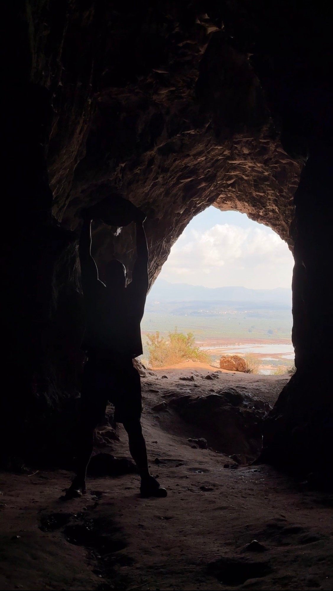 Silhouette lifting a rock overhead inside Nestor's Cave, with the bright opening and the lagoon behind