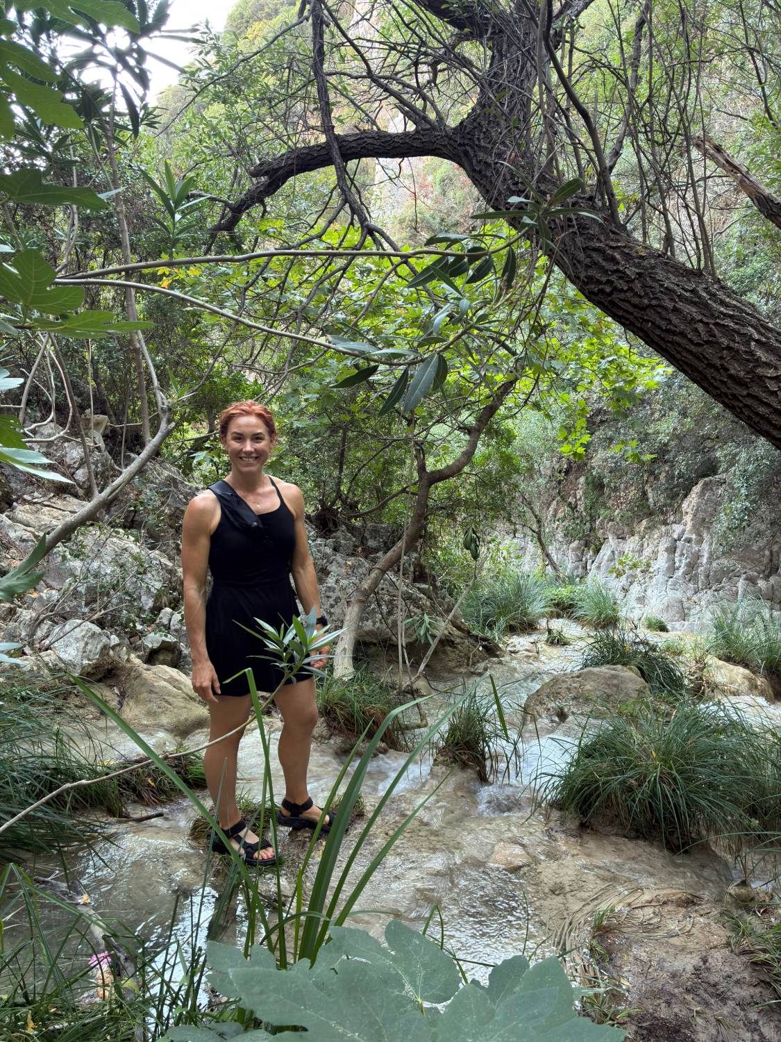 Kristine standing on the rocks of the stream at the very first cascades of Polylimnio, under the trees