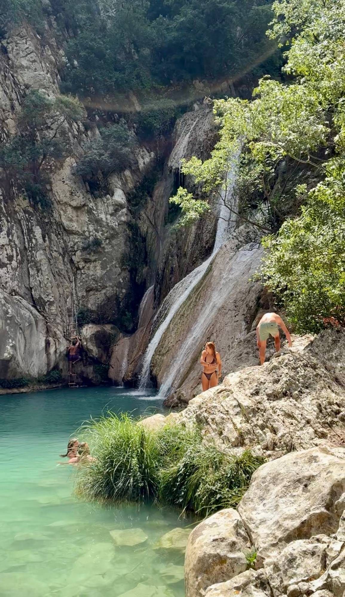 The tall waterfall at Lake Kadoula pouring down a rock face into a turquoise pool, with swimmers and a rope ladder
